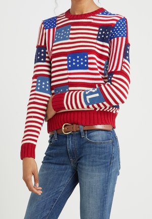Femme portant un pull en maille rouge, blanc et bleu avec des écussons du drapeau américain, associé à un jean bleu et une ceinture marron.