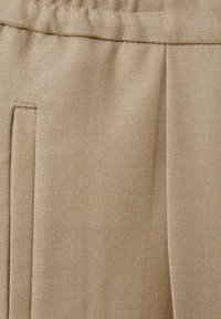 Tejido beige con un sutil patrón de espiga, que presenta una cinturilla y pliegues, mostrando una textura suave y un diseño estructurado.