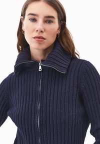 Merininauha neule korkealla kauluksella ja keskellä vetoketju; teksturoitu kangas, jossa on pystysuorat raidat ja tiukka istuvuus.