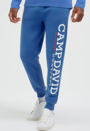 Man wearing blue sweatpants with "CAMP DAVID" and coordinates printed in white and red on the right leg, paired with white sneakers.