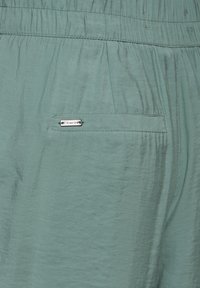 Helles teal-farbenes Gewebe mit einer weichen, leicht gekräuselten Textur. Verfügt über einen elastischen Bund und ein kleines, metallisches Markenetikett an einer Hintertasche.