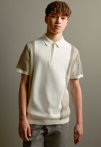 Īsroku polo krekls krēmkrāsā ar bēšām ribveida detalizācijām. Savukārt kakla daļā ir rāvējslēdzējs, un audums ir mīksts un adīts. Vienkārša muguras daļa, brīvs piegriezums.