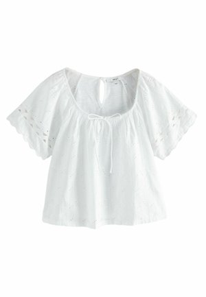 Femme portant un chemisier blanc à manches courtes avec broderie anglaise et bords festonnés, associé à un short en denim avec ourlet roulé, marchant en plein air.