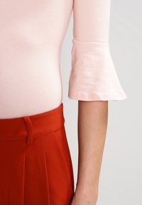 Svetlé růžové upnuté tričko s rozšířenými tříčtvrtečními rukávy spárované s texturovanými červenými kalhotami s vysokým pasem, které mají viditelnou poutko na pásek.