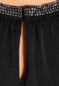 Svart tygöverdel med en pärlad krage prydd med klara, runda pärlor och ett centralt nyckelhålssnitt på baksidan. Mjuk textur.