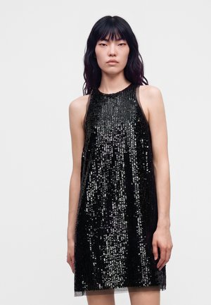 Un mannequin se tient devant un fond uni et clair, vêtu d’une robe noire sans manches couverte de sequins scintillants.