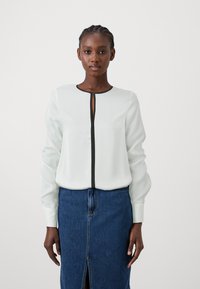 Bluză albă cu mâneci lungi, dotată cu o închidere cu nasturi negri și un tiv la gât, asortată cu o fustă din denim albastru, cu o fanta în față.