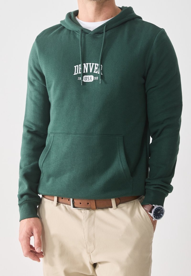 Zielona bluza z kapturem, z kieszonką z przodu, z białą grafiką „DENVER USA 98”. W zestawie z jasnymi spodniami i brązowym paskiem.