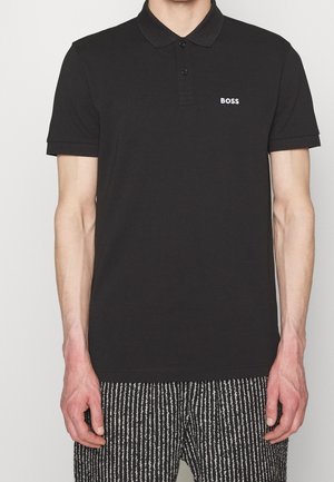 Mand iført en sort polo med "BOSS" logo på brystet og sort-hvide stribede bukser, stående med armene ned langs siderne.