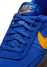 Kék velúr sneaker sárga Nike pálcával. Kék szövetfűzőkkel és párnázott nyelvvel rendelkezik. Texturált felület és kontrasztos anyagok jellemzik.