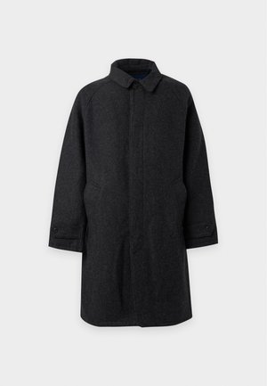 Mann trägt dunkelgrauen knöpfbaren Mantel, schwarzes Hemd, lila Hose und schwarze Schuhe und steht mit einer Hand in der Tasche vor schlichten Hintergrund.