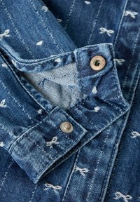 Cuello de chaqueta de mezclilla con un patrón de rayas azules y blancas, con broches de metal, tejido texturizado y sutiles detalles de costura.