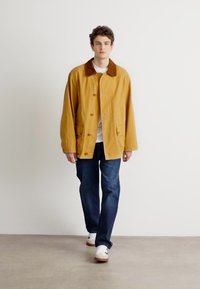 Giacca gialla leggera con colletto marrone, bottoni frontali e due tasche, abbinata a jeans scuri e sneakers bianche. Design semplice.