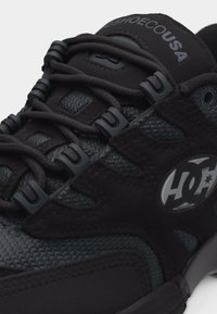 Κοντινό πλάνο μαύρου και σκουρόγκρι αθλητικού παπουτσιού με στρογγυλά κορδόνια, πλαστικούς θηλιές για κορδόνια, υφασμάτινη υφή και λευκό λογότυπο "DC" στο πλάι.