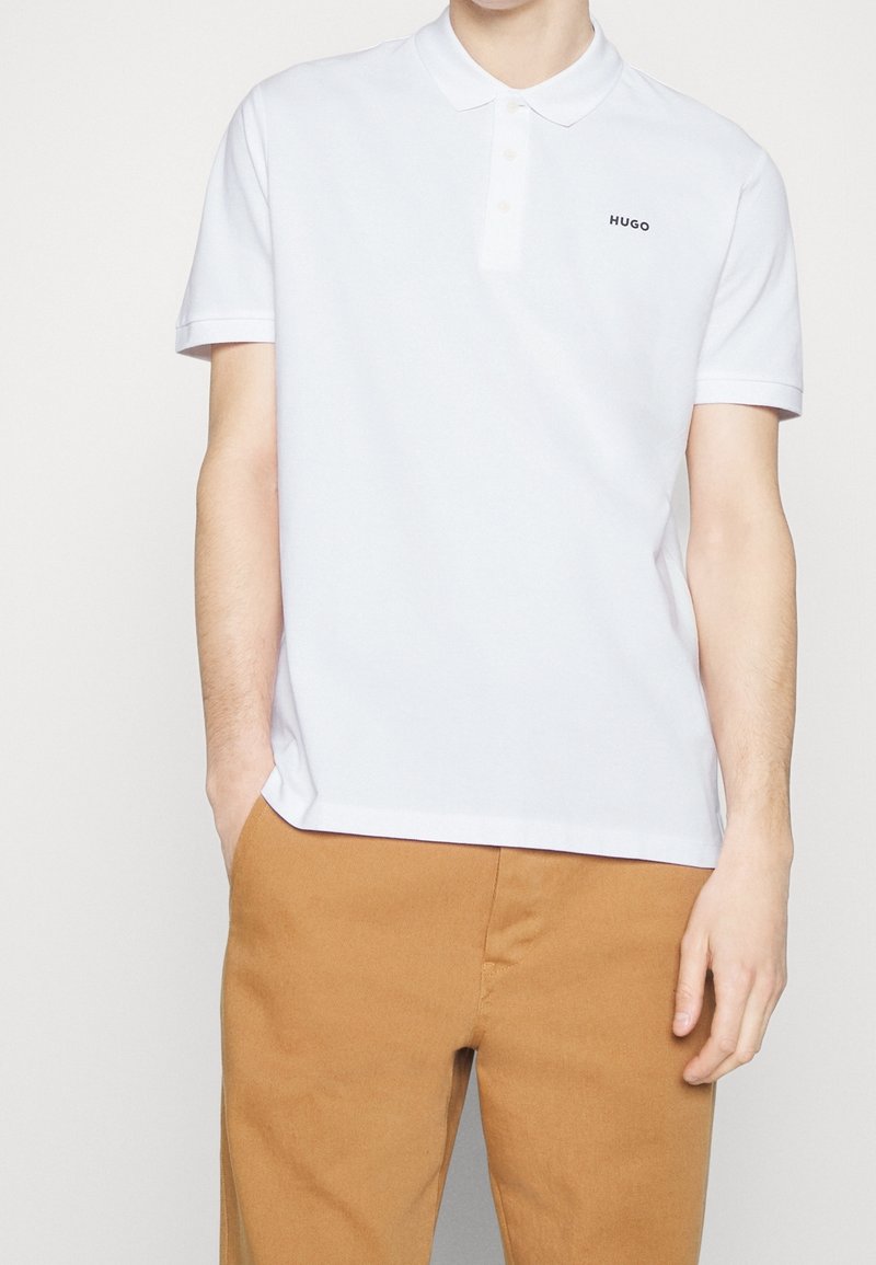 Homme portant un polo blanc à manches courtes avec le logo "HUGO" et un pantalon beige, debout avec une main dans la poche devant un fond uni.