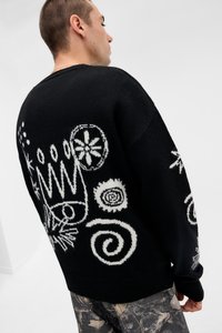 Μαύρο πλεκτό πουλόβερ με λευκά αφηρημένα σχέδια, συμπεριλαμβανομένου ενός στέμματος και σπειρών. Το ύφασμα φαίνεται υφασμένο με χαλαρή εφαρμογή.