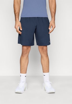 Mand iført marineblå træningsshorts, hvide Nike-strømper og hvide sneakers, stående mod en ensfarvet lys baggrund.