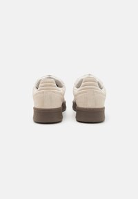 Beige suede sneakers with a soft texture, low-profile design, padded collar, and brown rubber outsole. Back view highlights stitching details.