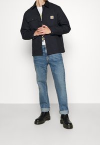 Dunkelblaues Arbeitsjackett mit Taschen, schwarzem Kragen, getragen über einem weißen Grafik-T-Shirt und hellblauen Jeans, kombiniert mit schwarzen Stiefeln.