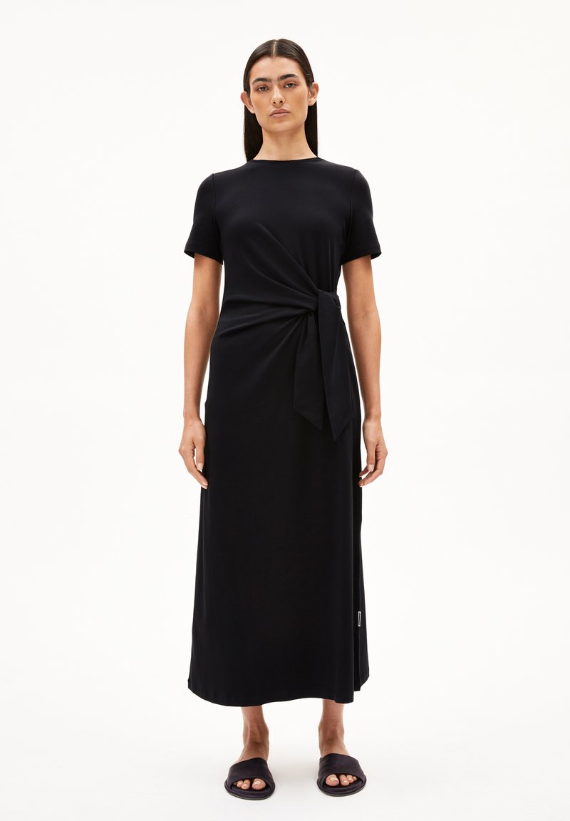Schwarzes, knielanges Kleid mit kurzen Ärmeln, gedrehten Frontdetails, weichem Stoff und relaxter Passform, kombiniert mit einfachen schwarzen Rutsch-Sandalen.