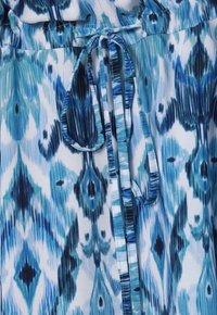 Modrý a bílý vzorovaný textil s uzávěrem na zavázání, který má design ikat, měkkou strukturu a různé odstíny modré a černé.