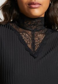 Haut noir à col montant avec un col en dentelle à motifs floraux et une texture côtelée, créant une silhouette ajustée.