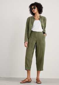 Olivinvihreä pellavapuku, jossa on napit, yhdistettynä samanvärisiin lyhyisiin housuihin. Valkoinen paita alla ja ruskeat raskaansarakintaiset sandaalit. Mustat aurinkolasit.