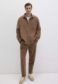 Braunes Cordhemd mit einer Tasche, kombiniert mit passenden braunen Hosen. Helle Sneakers runden das Outfit ab. Einfaches und strukturiertes Design.