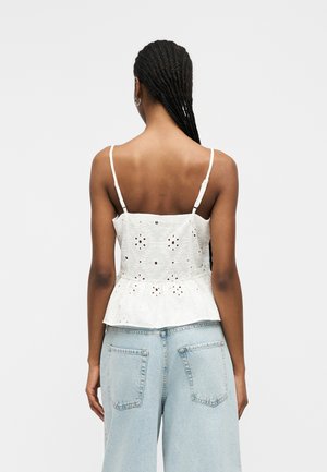 Kvinne med lange fletter iført en hvit ermeløs øyelet-top med justerbare stropper og lyseblå høytaljede jeans, vist bakfra.