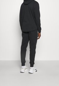Persona che indossa un felpa nera con cappuccio e pantaloni jogger neri con scarpe da ginnastica bianche, in piedi contro uno sfondo chiaro uniforme, vista posteriore.
