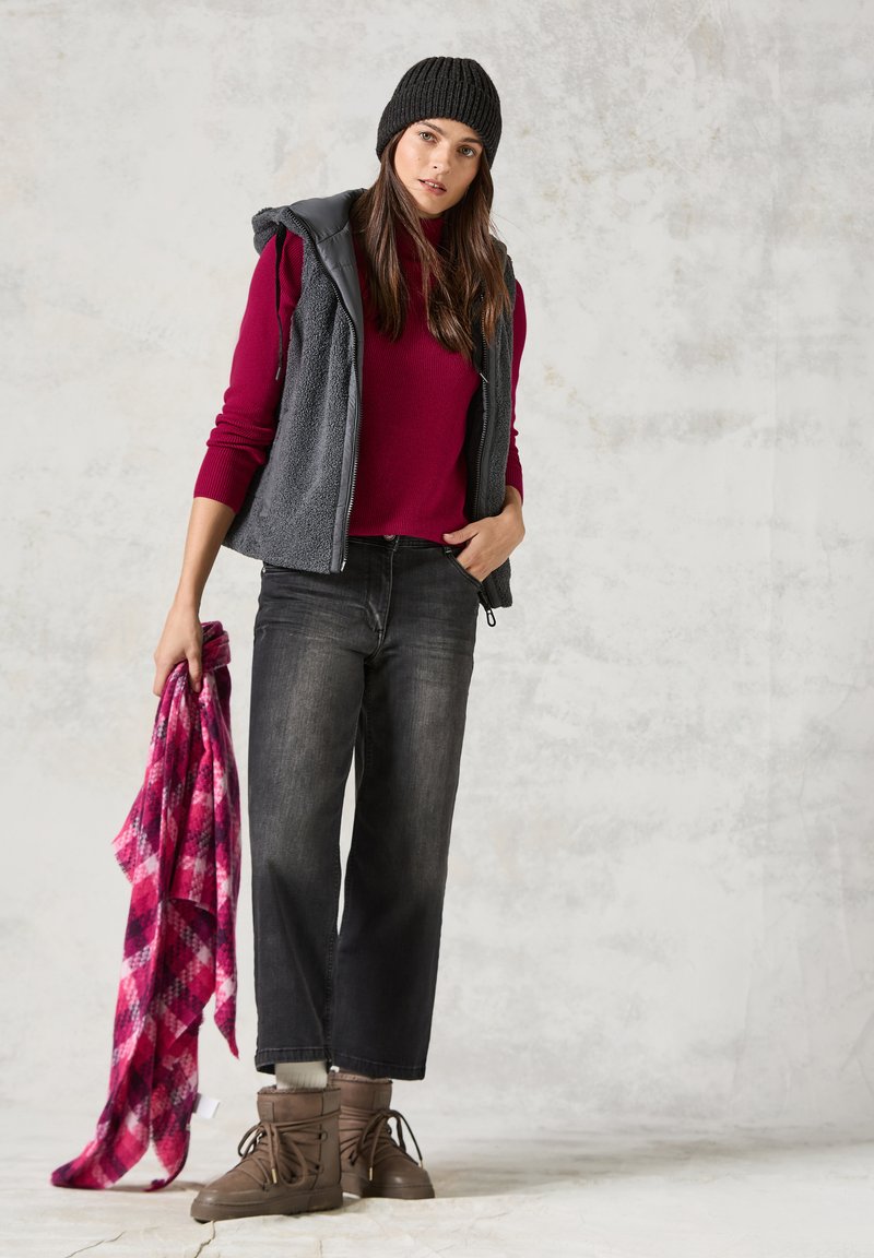 Grau Fleece-Weste über einem burgunderfarbenen Pullover, schwarze weite Jeans und braune Schnürstiefel; hält einen rosa karierten Schal.