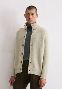 Jasnobeżowy sweter z dzianiny z prążkowanym kołnierzem, zapięciem na guziki z przodu i długimi rękawami. Noszony na ciemnym golfie, widoczna jest fakturowana tkanina.