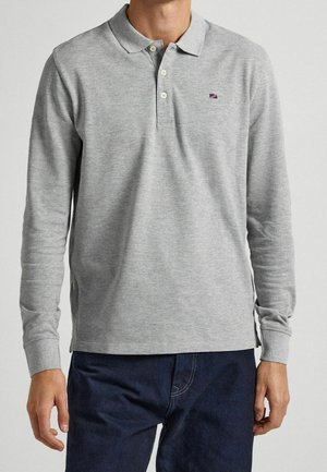 Uomo che indossa una polo grigio chiaro a maniche lunghe con tre bottoni e pantaloni blu scuro su uno sfondo semplice.