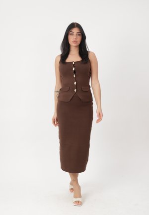 Frau mit langen dunklen Haaren, die ein ärmelloses braunes, geknöpftes Oberteil und einen passenden Midi-Rock trägt, geht vorwärts in weißen offenen Schuhen mit Absatz.