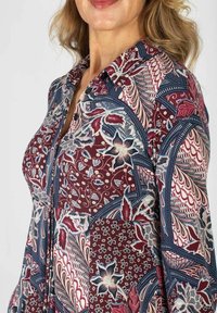 Kobieta w koszuli na guziki z wielokolorowym wzorem kwiatowym i abstrakcyjnym w odcieniach burgundu, granatu, niebieskiego i kremowego.