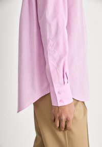 Camicia a maniche lunghe rosa chiaro con vestibilità sartoriale, caratterizzata da polsini con bottone e orlo curvo. Il materiale appare morbido e liscio.
