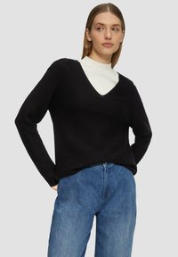 Zwarte V-hals trui met lange mouwen, gestructureerde breiwijze, en eroverheen gedragen een witte kraagloze trui, gecombineerd met lichtblauwe denim jeans.