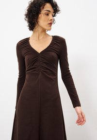 Braun langärmliges Kleid aus weichem Stoff, mit einem gerafften V-Ausschnitt und einer fließenden A-Linien-Silhouette. Glatte Textur, taillierter Oberkörper.