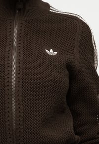 Braune Strickjacke mit Reißverschluss, Adidas-Logo auf der Brust und weißen Streifen entlang der Schulter und des Ärmels.