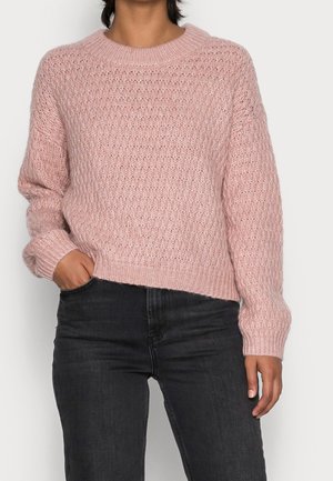 Person trägt einen texturierten, hellrosa Strickpullover mit langen Ärmeln und schwarze High-Waist-Jeans, eine Hand in der Tasche, steht vor neutralem Hintergrund.