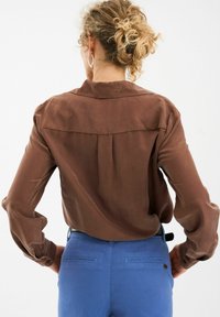 Bluză din mătase maro cu mâneci lungi și detaliu la spate, asortată cu pantaloni blue cu o croială ajustată și buzunare la spate.