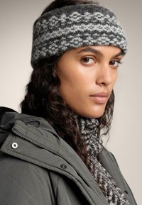 Pletená čelenka v sivej farbe s bielymi kvetinovými vzormi, skombinovaná so sivým svetrom s vysokým golierom a zelenou prešívanou bundou. Viditeľné textúrové detaily.