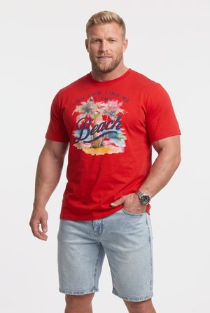 Uomo muscoloso con capelli biondi corti che indossa una T-shirt rossa con grafica "Beach" e pantaloncini in denim chiari, una mano in tasca, sfondo chiaro.