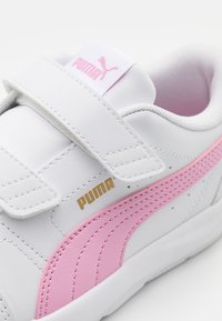 Sneaker sintetico bianco con accenti rosa, dotato di una fascia in Velcro, dettagli testurizzati e marchio dorato. Superficie liscia e forma arrotondata.