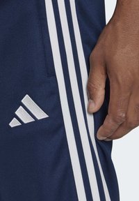Marinefarbene Sportshorts mit weißen Drei-Streifen-Akzenten, die ein gesticktes weißes Logo am linken Bein aufweisen, sowie eine Nahaufnahme einer Hand, die den Stoff greift.