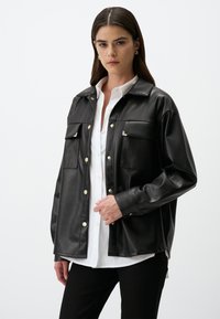 Giacca camicia in similpelle nera con bottoni a pressione dorati e due tasche sul petto. Maniche lunghe con una vestibilità rilassata, indossata sopra una camicia bianca.