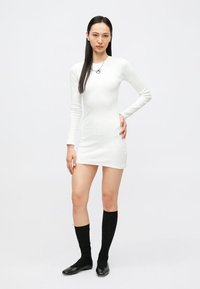 Biała prążkowana sukienka z długim rękawem o dopasowanym kroju, zestawiona z czarnymi podkolanówkami i czarnymi płaskimi butami. Minimalistyczny design.