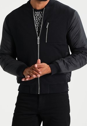 Homme portant une veste bomber noire avec une fermeture éclair sur le devant et une poche poitrine zippée, les mains croisées à la taille, avec une chemise à motifs en dessous.