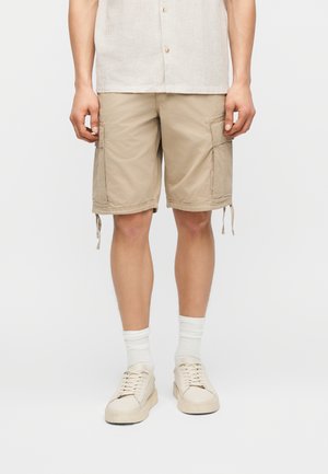 Άνδρας που φοράει μπεζ cargo βερμούδες, λευκές κάλτσες και μπεζ αθλητικά παπούτσια, στέκεται μπροστά σε ένα απλό ανοιχτό γκρι φόντο.