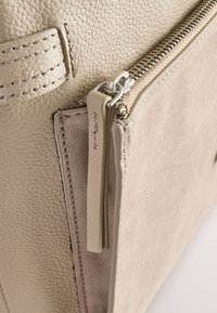 Detaliu al unei genți din piele și velur beige care prezintă o suprafață texturată, fermoar vizibil și un accent moale de tip etichetă. Include margini cusute și accesorii netede.